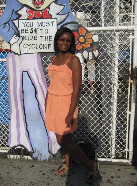 Dark skinned Caribbean & African American woman in Coney Island, Brooklyn, New York, USA wearing a orange dress in front of a sign that reads You must be 54 inches to ride the Cyclone
